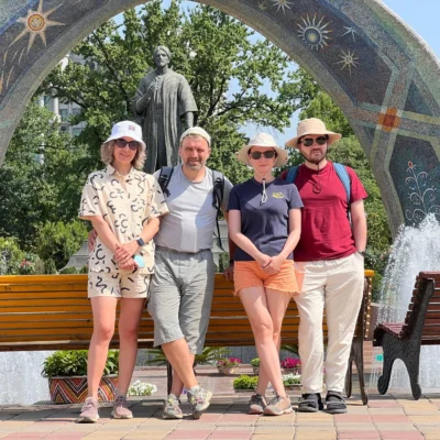 Quattro amici sono in piedi su una panchina di legno di fronte a una statua e a un arco a mosaico, e sorridono alla macchina fotografica.