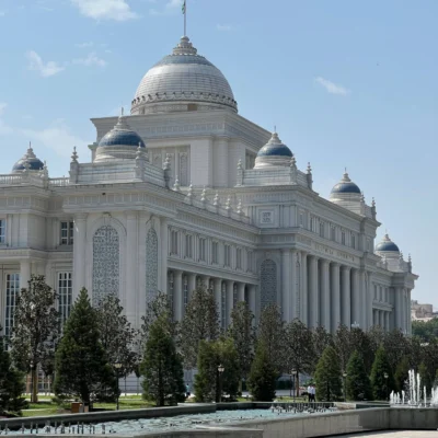 White neoclassical building with blue-domed cupolas and ornate details, set behind trees and a water feature in the foreground.