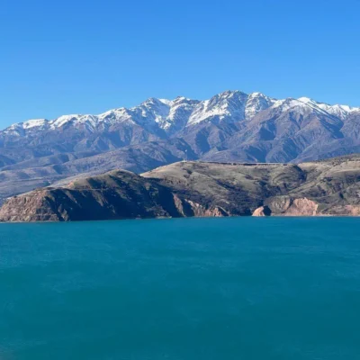 Dietro una costa frastagliata e una baia turchese, sotto un cielo azzurro e limpido, si ergono montagne innevate.”