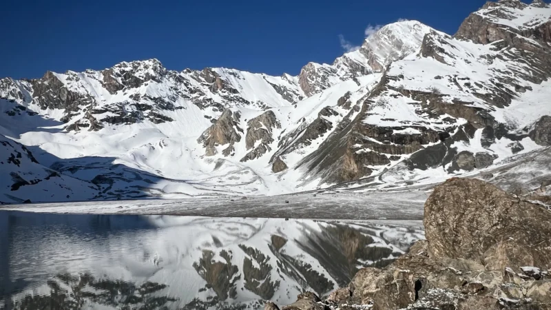 Montagne frastagliate e innevate sotto un cielo azzurro e limpido, con un lago immobile e riflettente in primo piano.