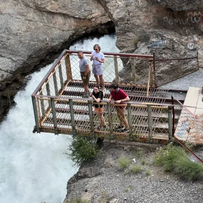 Four people stand on a rusted metal viewing platform over a fast-flowing waterfall in a rocky canyon with graffiti on the walls behind them.