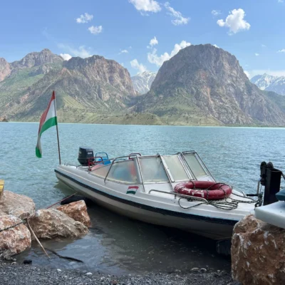 White motorboat moored at a rocky lakeshore with a flag, calm blue water, and a jagged mountain range beyond.