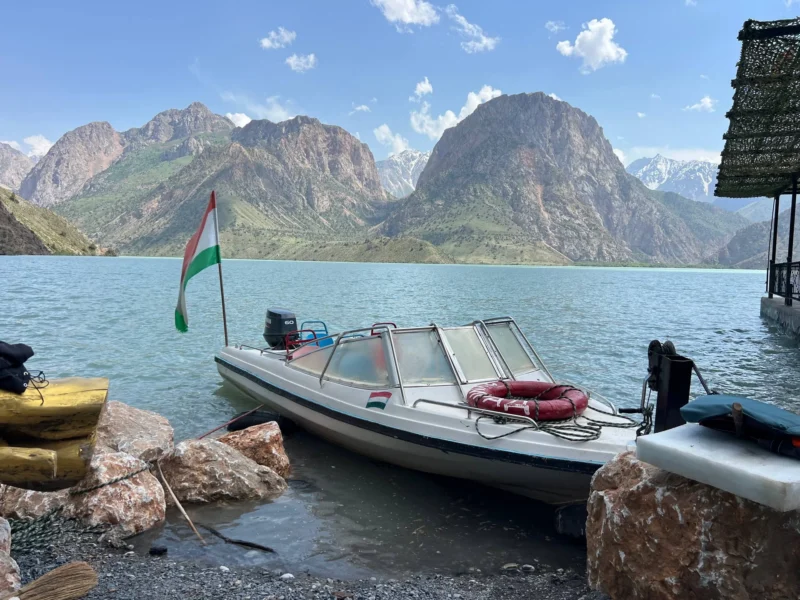 Un motoscafo bianco ormeggiato sulla riva rocciosa di un lago, con una bandiera al vento, acque calme e blu e una catena montuosa frastagliata sullo sfondo.