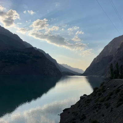 Calm mountain valley with a reflective lake between steep rocky slopes and a clear blue sky with scattered clouds in the background.