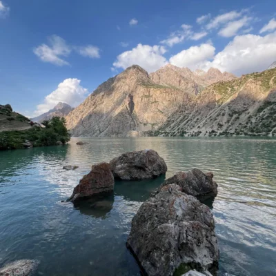 Lago turchese con rocce in primo piano e ripide montagne rocciose sotto un cielo azzurro limpido con nuvole.