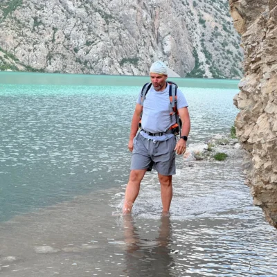 Un escursionista con maglietta grigia e zaino in spalla cammina lungo la riva rocciosa di un lago alpino turchese, visibile tra le scogliere.