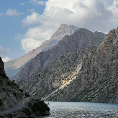 Mountain range with steep rocky cliffs beside a calm lake and blue sky with white clouds